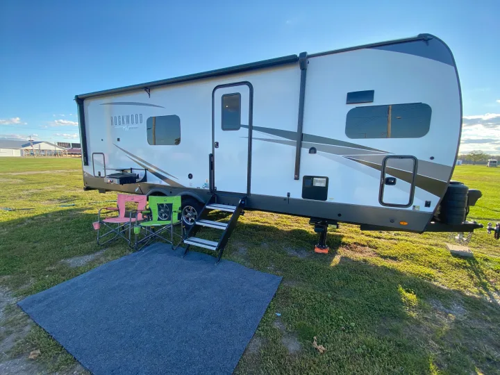 A modern Rockwood travel trailer (RV rental) at a sunny campsite, with an awning, steps, chairs, and a mat.. Forest River Rockwood Signature 2024