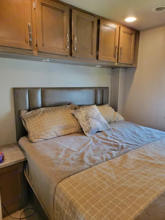 Inside an RV rental: a cozy bedroom with a large bed, brown headboard, and wood-paneled overhead cabinets.. Entegra Coach Vision 2019