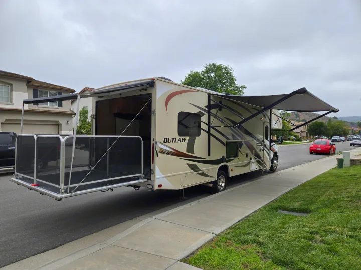 This rv rental, a beige Outlaw toy hauler, is parked with its rear ramp and side awning extended on a residential street.. Thor Outlaw 2016