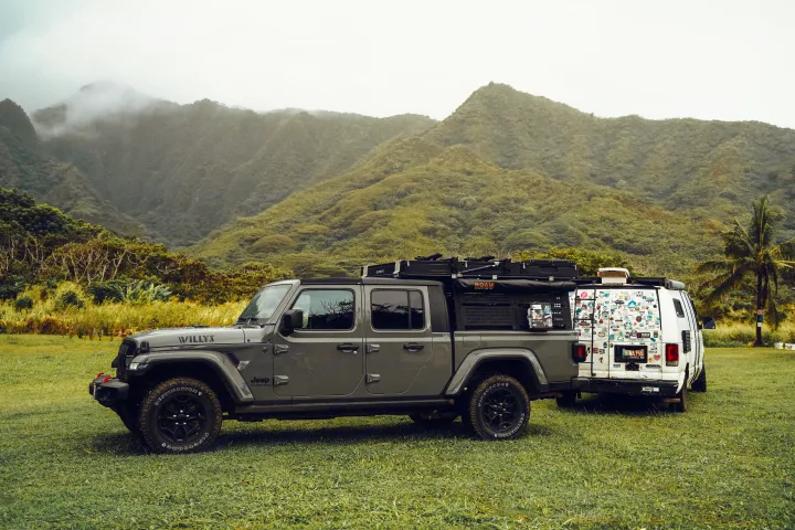 A gray Jeep Gladiator pickup and a white van RV, sticker-covered, parked in a grassy field with misty mountains. Ideal for RV rental trips.. Jeep Gladiator 2022