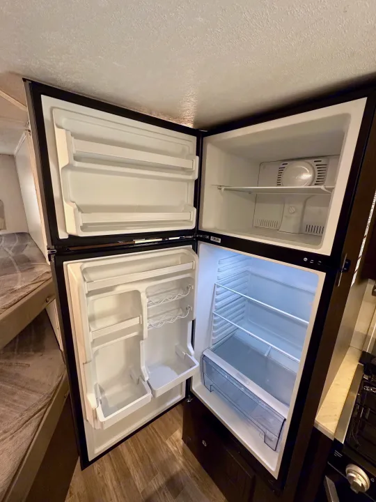 Open, empty two-door RV rental fridge with white interior, ready for groceries. Freezer on top, main compartment below.. Forest River Wildwood X-Lite 2020