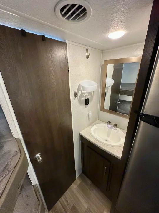 Compact RV rental bathroom featuring a vanity, sink, mirror, dark wood door, and a reflection of a bunk area.. Forest River Wildwood X-Lite 2020
