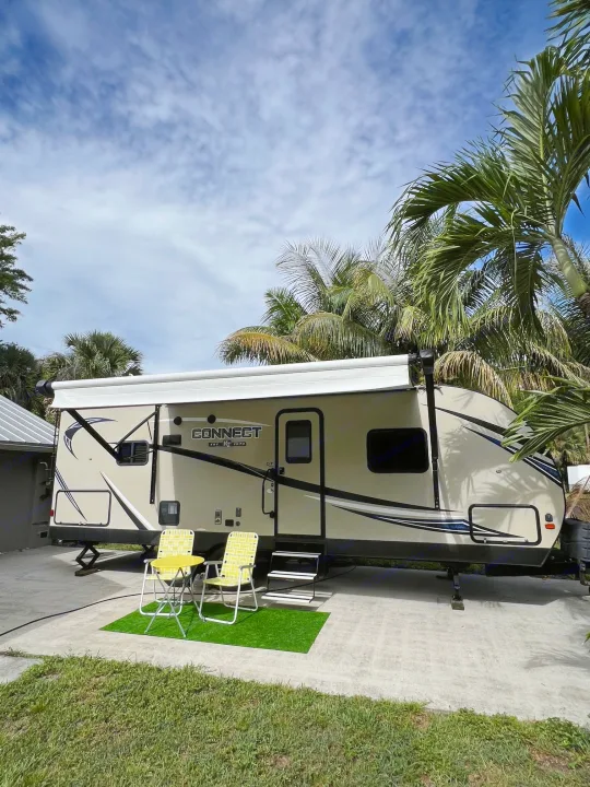 Relaxing RV rental setup: A tan travel trailer with awning, yellow chairs & table, surrounded by palm trees.. KZ Connect RLK 24 2019