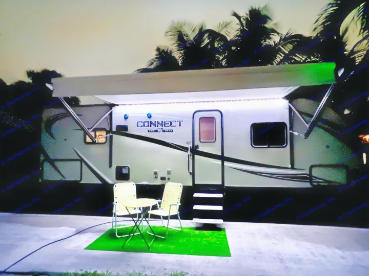 Cozy night at an RV rental campsite. Illuminated RV awning, outdoor seating, and palm trees create a welcoming scene.. KZ Connect RLK 24 2019