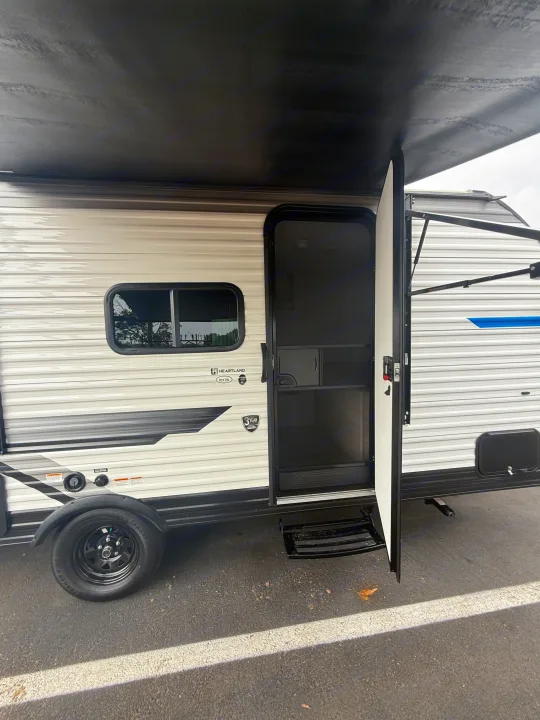 This rv rental is a white Heartland travel trailer with open door, screen, and steps, under an extended black awning.. Heartland Pioneer 2025