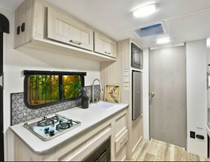 Step inside this compact RV rental kitchen featuring modern light wood, built-in sink, stovetop, microwave, and forest view.. Heartland Pioneer 2025