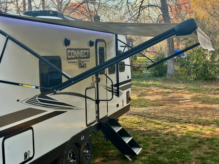 A white travel trailer RV rental with an extended awning, featuring bright LED lights, parked in a sunny, grassy spot.. KZ Connect SE 2024