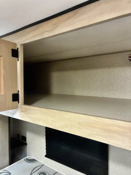 Empty light wood overhead cabinet with black hinges in an RV rental, showcasing interior storage above a bed and window.. KZ Connect SE 2024