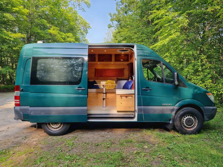 Green RV rental van parked in a forest, showcasing its custom wood interior through the open side door.. Dodge Sprinter Van 2008