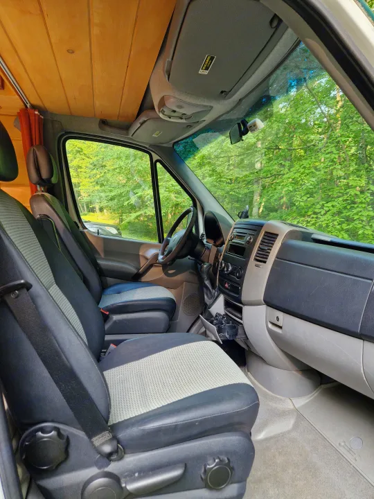 Front cabin of an RV rental: driver and passenger seats, dashboard, wooden ceiling, and green trees outside.. Dodge Sprinter Van 2008