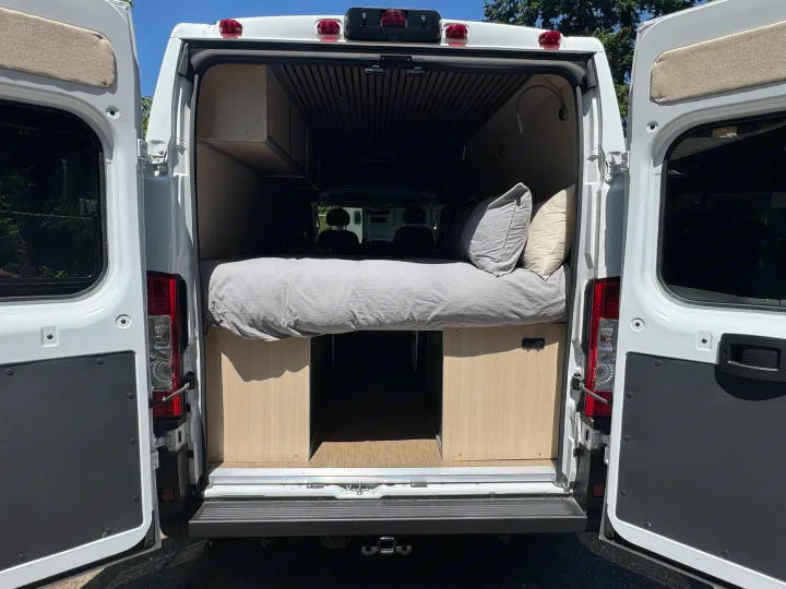 Rear view of an RV rental camper van, revealing its cozy bed, pillows, and built-in wooden storage. Ready for adventure.. Dodge B Van 2023