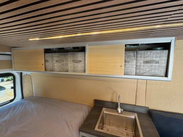 Spacious RV rental interior with bed, kitchen sink, overhead storage, and modern slat ceiling with lighting.. Dodge B Van 2023