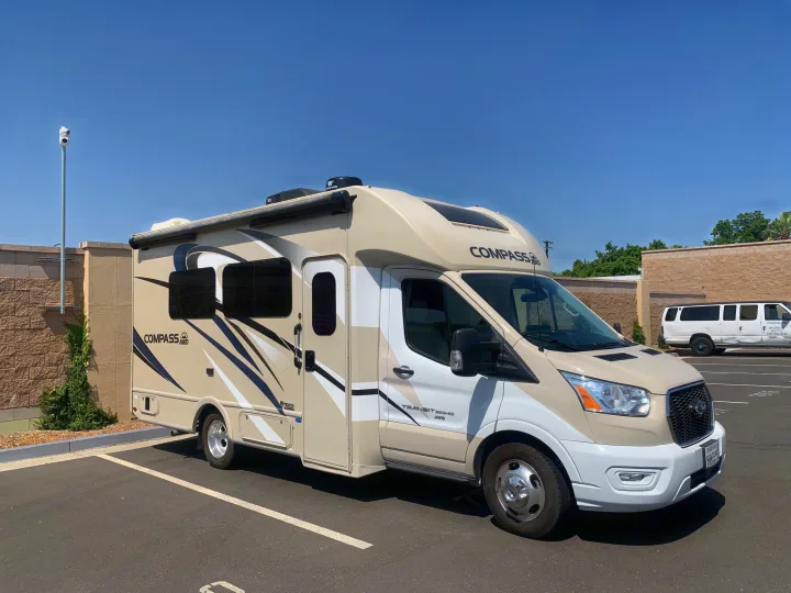 A tan and white Thor Compass AWD rv rental, a Class C RV on a Ford Transit chassis, parked in a paved lot on a sunny day.. Thor Compass 2021
