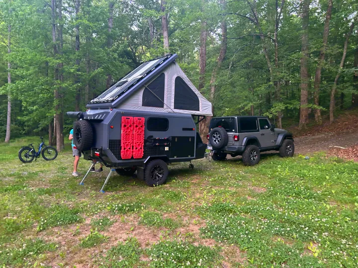 RV rental: Grey off-road camper trailer with rooftop tent & solar panels, hitched to a Jeep in a wooded campsite.. Zoomies Campers Spirit 2025