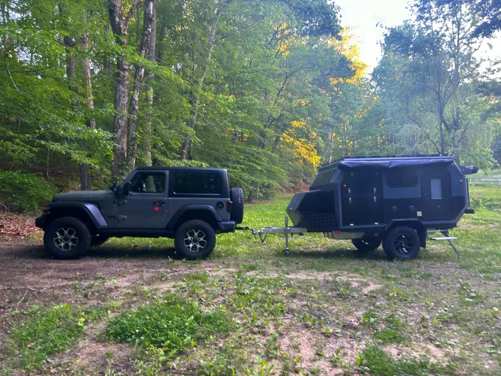 Dark grey Jeep towing a black off-road RV rental trailer in a lush forest clearing.. Zoomies Campers Spirit 2025