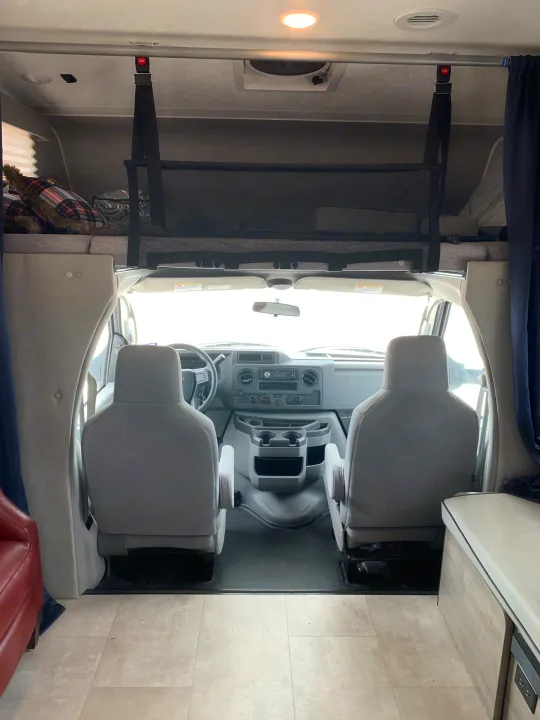View into an RV rental cockpit from the living area, featuring the dashboard, front seats, and an overhead bunk with safety net.. Coachmen Freelander 2021