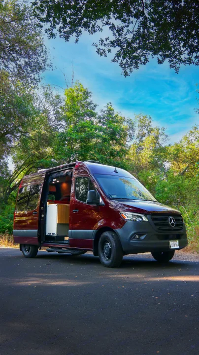 Maroon Mercedes Sprinter RV rental parked on a road, side door open to reveal a custom interior, surrounded by green trees.. Mercedes-Benz Sprinter 2025