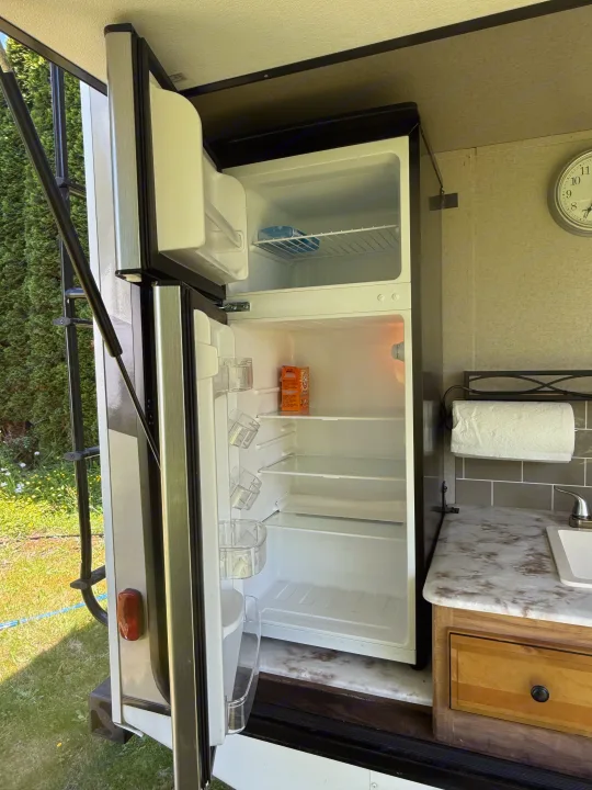 An open RV rental refrigerator in an outdoor kitchen area with a counter, sink, and paper towels, next to an exterior ladder.. Coachmen Freedom Express Ultra Lite Maple Leaf Edition 2019