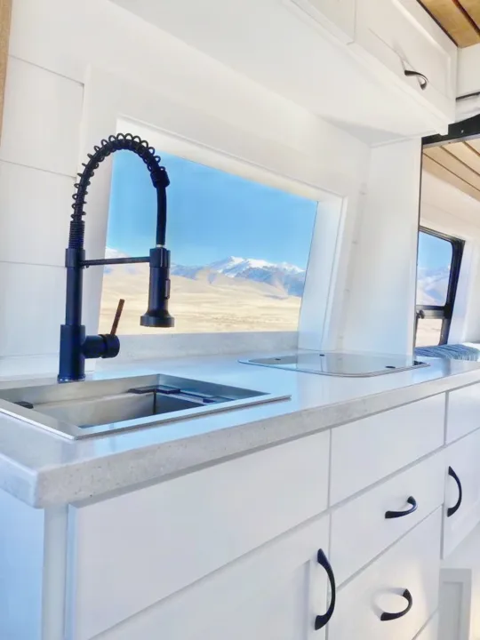 Modern RV rental kitchen with white cabinets, sleek black faucet, compact cooktop, and amazing mountain views from large windows.. Custom Crafted Vans Family Van 2019