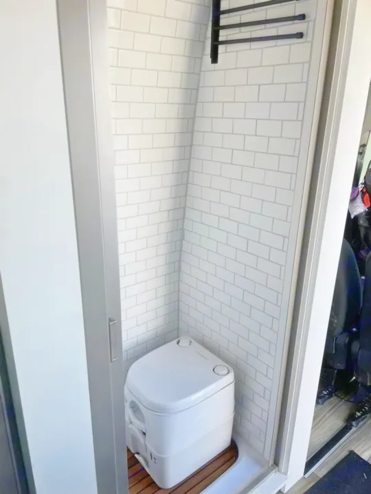 Inside an RV rental: a compact, tiled wet bath featuring a composting toilet on a wooden mat and a towel rack.. Custom Crafted Vans Family Van 2019