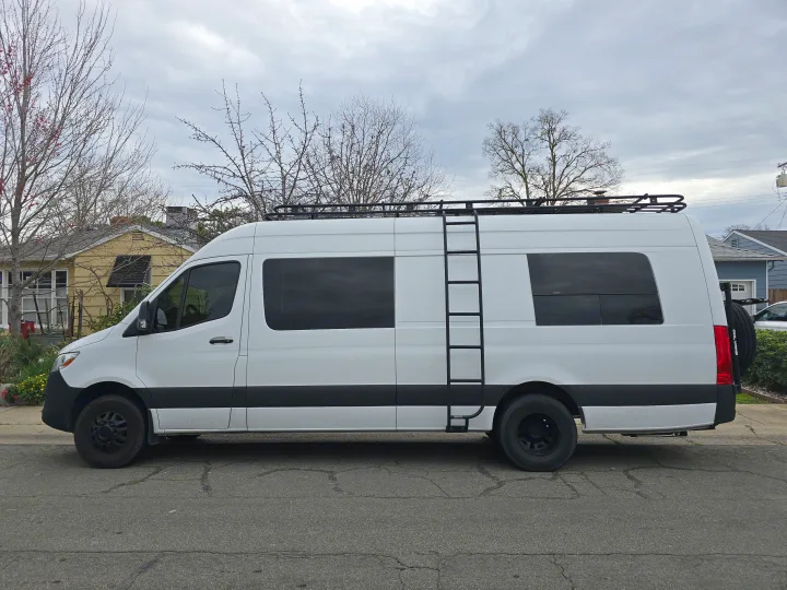 White Mercedes Sprinter camper van for RV rental, featuring a roof rack and side ladder.. Custom Crafted Vans Family Van 2019