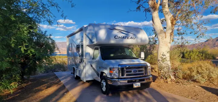 White Chateau RV rental parked at a scenic campsite with mountains and a lake in the background under a blue sky.. Thor Motor Coach Chateau 2026