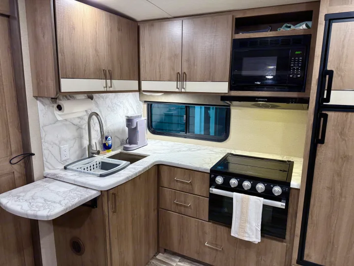 Modern RV rental kitchen with wood cabinets, marble-look counters, microwave, stove, sink, and coffee maker.. Grand Design Imagine 2021