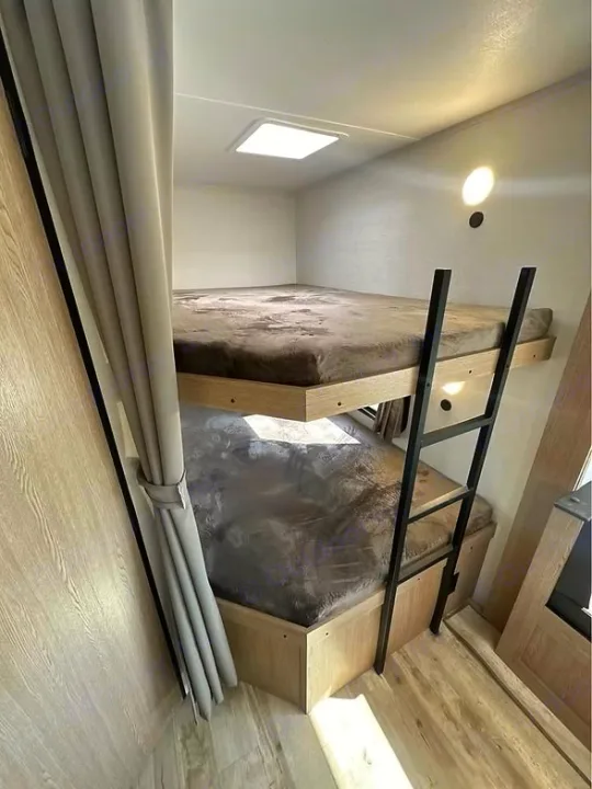 Comfortable bunk beds in a modern RV rental. This bright, compact interior features a ladder, light wood finishes, and a gray curtain.. Gulf Stream Amerilite 2025