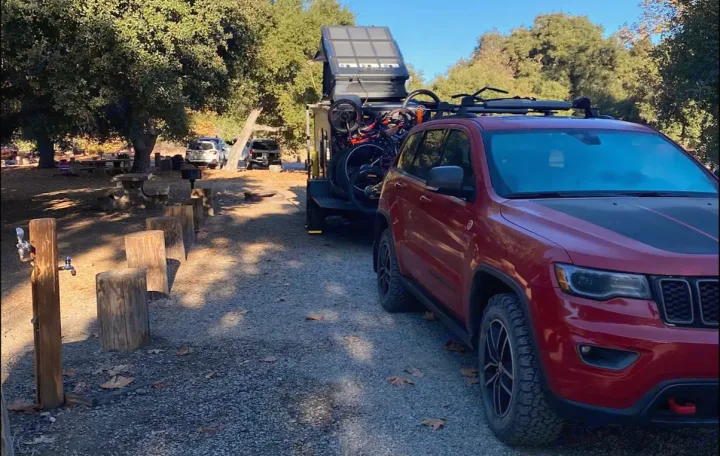 Red Jeep Grand Cherokee towing an adventure trailer with an RV rental rooftop tent and bikes at a sunny campground.. ITWO custom Pixtech Bearlander 20 2024