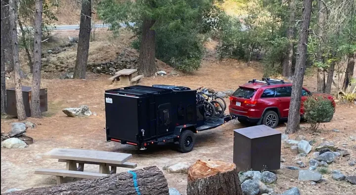 This overland camper, perfect for an RV rental, is towed by a red SUV with bikes at a rustic forest campsite.. ITWO custom Pixtech Bearlander 20 2024