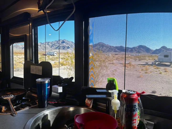 RV rental kitchen counter with a stove, sink, and a sunny desert view of mountains and other RVs through the windows.. Airstream Basecamp 2024