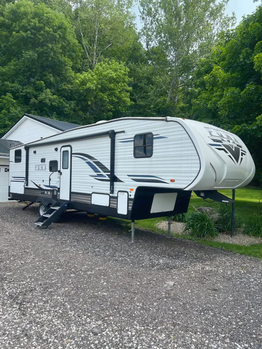 White Puma fifth-wheel RV rental parked on a gravel driveway, nestled beside a house with lush green trees.. Forest River Palomino Puma 2022