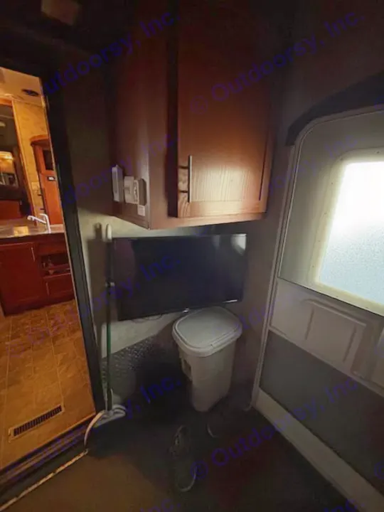 Dimly lit RV rental interior: overhead cabinets, TV, white trash bin, broom, and a glimpse of the kitchen.. Keystone RV Fuzion Toy Hauler 2009