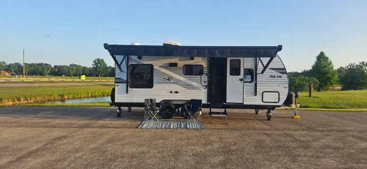 An RV rental, a travel trailer with its awning extended, chairs, and mat set up by a pond, ready for camping.. Jayco Jay Flight SLX 210 QB 2025