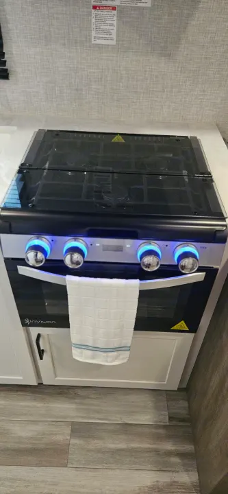 This RV rental kitchen boasts a modern, compact stove and oven, featuring illuminated blue control knobs, a glass top, and propane safety signage.. Jayco Jay Flight SLX 210 QB 2025