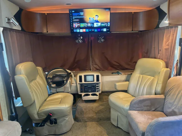Inside a comfy RV rental: beige cockpit seats, dash, and TV displaying streaming apps. Brown curtains add privacy for your travel.. Winnebago Sightseer 2009