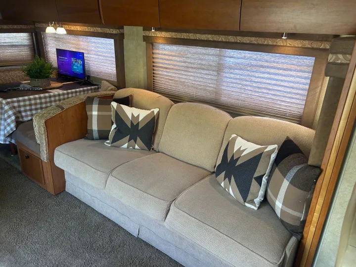 Cozy RV rental interior with a light-colored sofa, patterned pillows, and a dining area with a TV and plant.. Winnebago Sightseer 2009