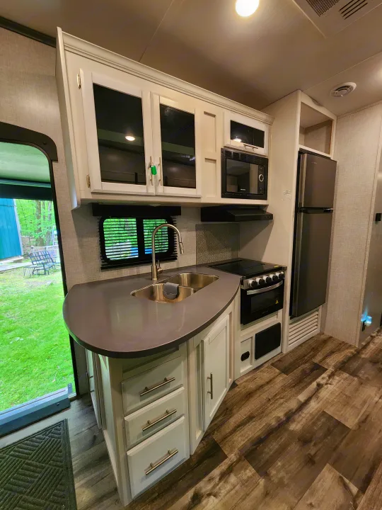 Modern RV rental kitchen: white cabinets, dark counters, sink, and appliances. Bright interior with view to green yard.. Crossroads RV Volante 2022