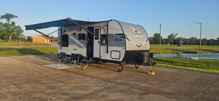 Jayco RV rental parked at a scenic campsite with an extended awning, chairs, and a rug. Perfect for camping beside a pond.. Jayco Jay Flight SLX 210 QB 2025