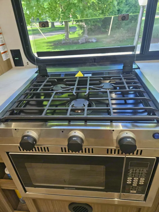 RV rental kitchen featuring a modern stainless steel gas stove with three burners and an integrated microwave oven.. Forest River 26Ice 2025