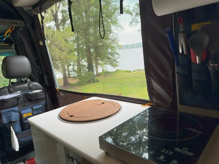 RV rental interior features a compact kitchen with an induction cooktop, cutting board, and serene lake view through a mesh screen.. Storyteller Overland Storyteller Overland Class B 2023