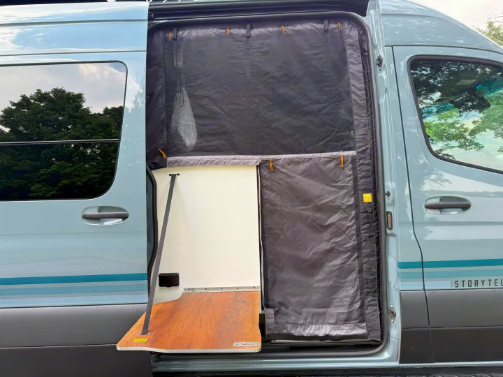Open side door of a light blue Storytel van, showcasing a pull-out table and protective mesh screen. Perfect for your rv rental.. Storyteller Overland Storyteller Overland Class B 2023