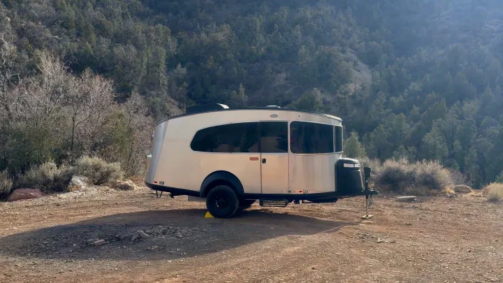Modern silver travel trailer RV rental at a remote campsite, nestled against tree-covered mountains.. Airstream Basecamp 2024