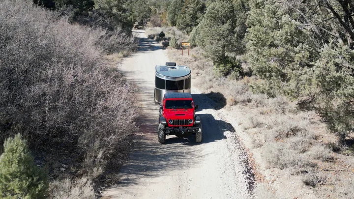 Red Jeep towing a silver RV rental on a dirt road through dry forests. Perfect for your off-grid adventure.. Airstream Basecamp 2024