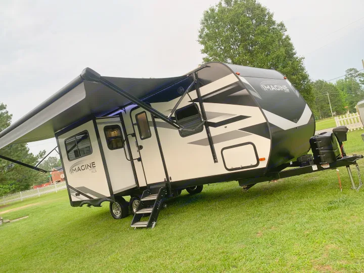 White & black Imagine travel trailer on green grass, awning extended with lights. Ideal for an RV rental adventure.. Grand Design Imagine XLS 2022