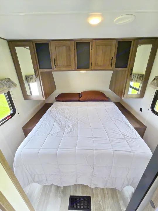 Inside a comfortable RV rental bedroom, featuring a queen bed, wooden storage cabinets, and side windows with green views.. Avenger Avenger Trailer 2021