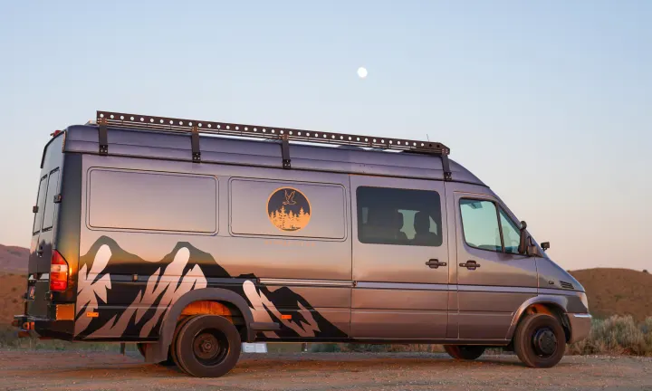 This grey camper van, ideal for RV rental adventures, features mountain graphics and a roof rack under a soft, moonlit sky.. Dodge Sprinter Van 2005