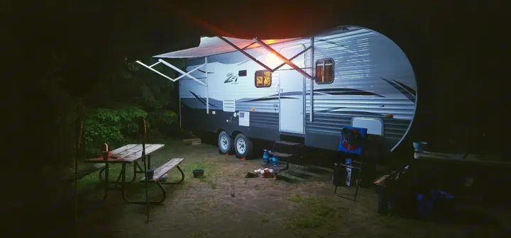 Nighttime RV rental camping scene: a travel trailer with an illuminated awning, picnic table, and gear under a dark sky.. Crossroads RV Z-1 2015