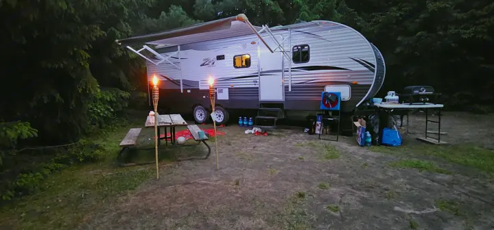 Evening at an RV rental campsite: a travel trailer, picnic table, and grill are illuminated by tiki torches under dark trees.. Crossroads RV Z-1 2015