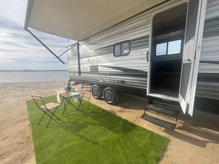 Waterfront RV rental setup: travel trailer with extended awning, outdoor turf, table, and chairs by the bay.. Forest River EVO 2021